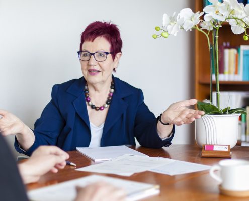 Die Anwältin sitzt am Besprechungstisch. Darauf liegen Unterlagen. Sie berät einen Mandanten, der nur von hinten zu sehnen ist.